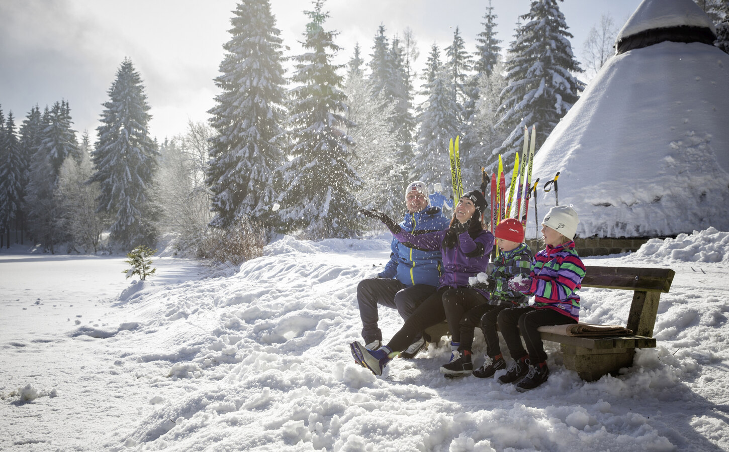 Eine Familie sitzt auf einer Holzbank in der verschneiten Landschaft.